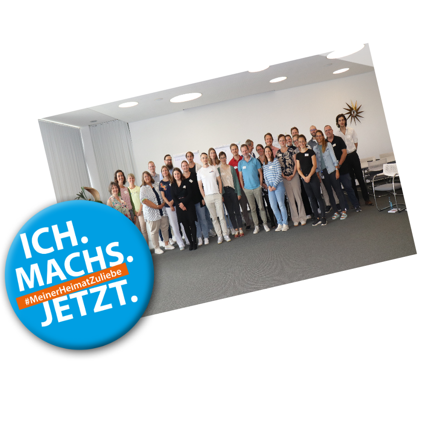 Blaues Logo mit Text: "Ich machs jetzt #MeinerHeimatZuliebe" auf einem Gruppenbild der Klimaschutzmanagerinnen und -managern in einem großen Raum